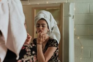Woman with towel around her head applying makeup in a bathroom mirror